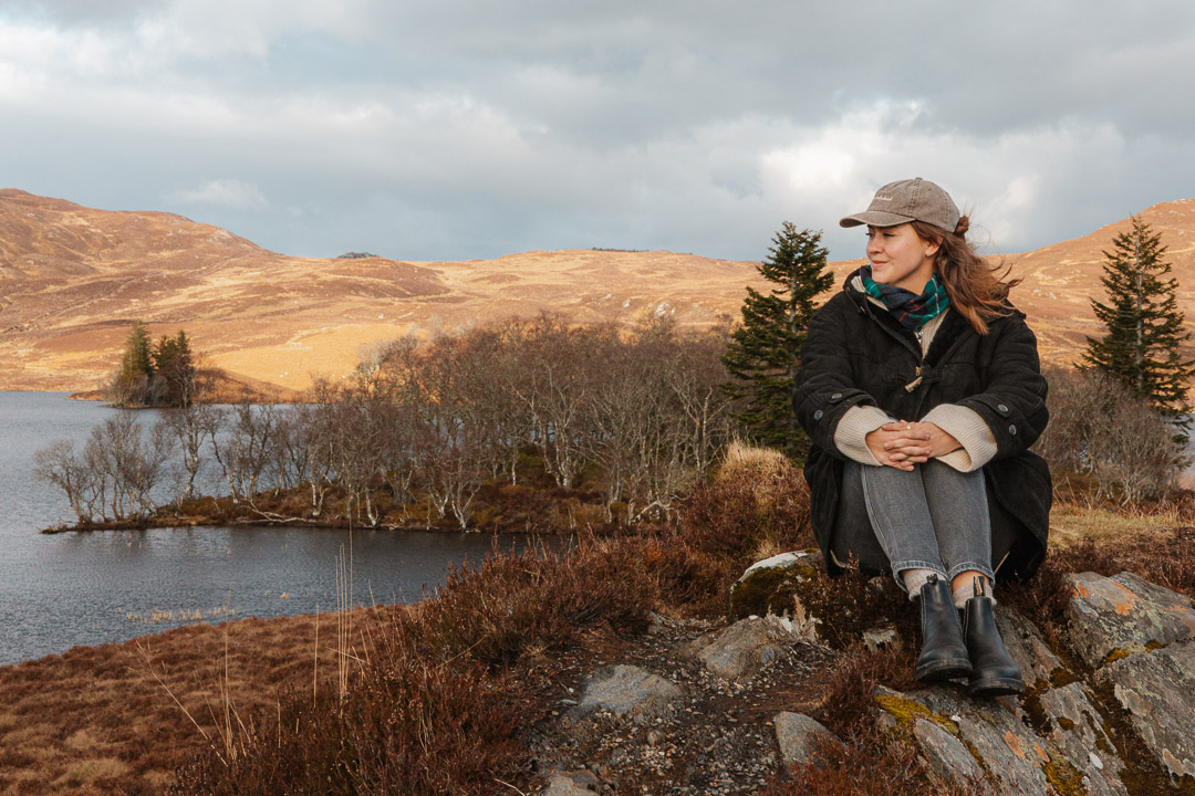 Frau sitzt in der Natur in Schottland, herbstliche Atmosphäre, im Hintergrund ein See mit einer Insel